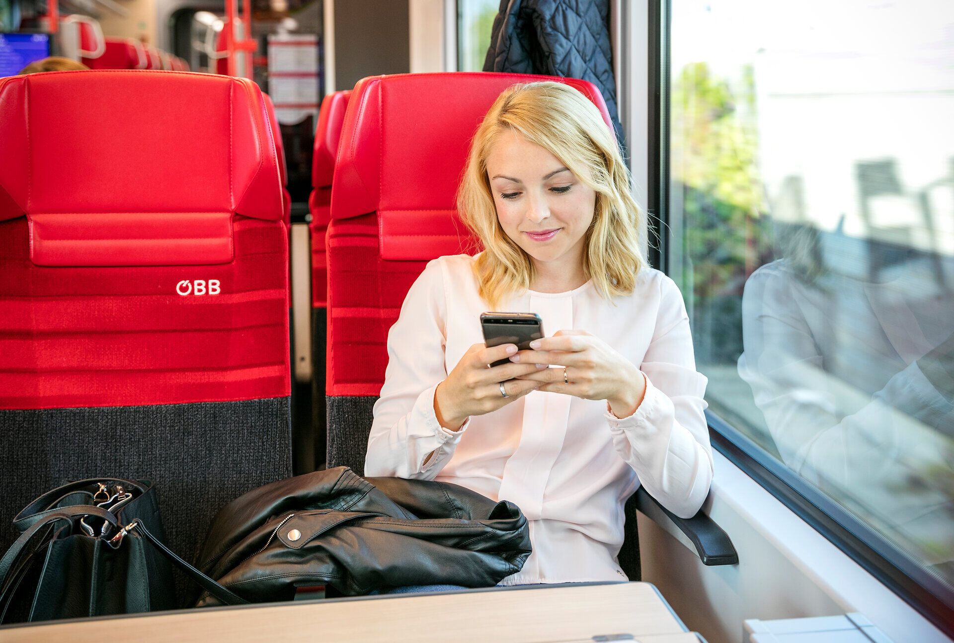 Eine junge Frau genießt die entspannte Atmosphäre während ihrer Zugfahrt. Umgeben von modernen, roten Sitzen, ist sie in ihr Smartphone vertieft und lässt sich von der vorbeiziehenden Landschaft inspirieren. Diese Reise verspricht unvergessliche Erlebnisse und neue Entdeckungen.