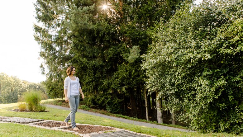 A woman walks barefoot on a green path in the spa gardens of Bad Sch&ouml;nau, surrounded by trees and sunlight.