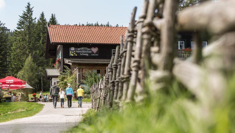 Drei Personen gehen auf einen Berggasthof zu, umgeben von einem Holzzaun und Bäumen.