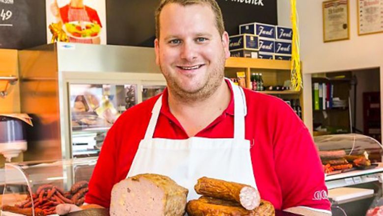 Ein Metzger in rotem Hemd und weißer Schürze hält verschiedene Wurstwaren in einer Fleischerei.