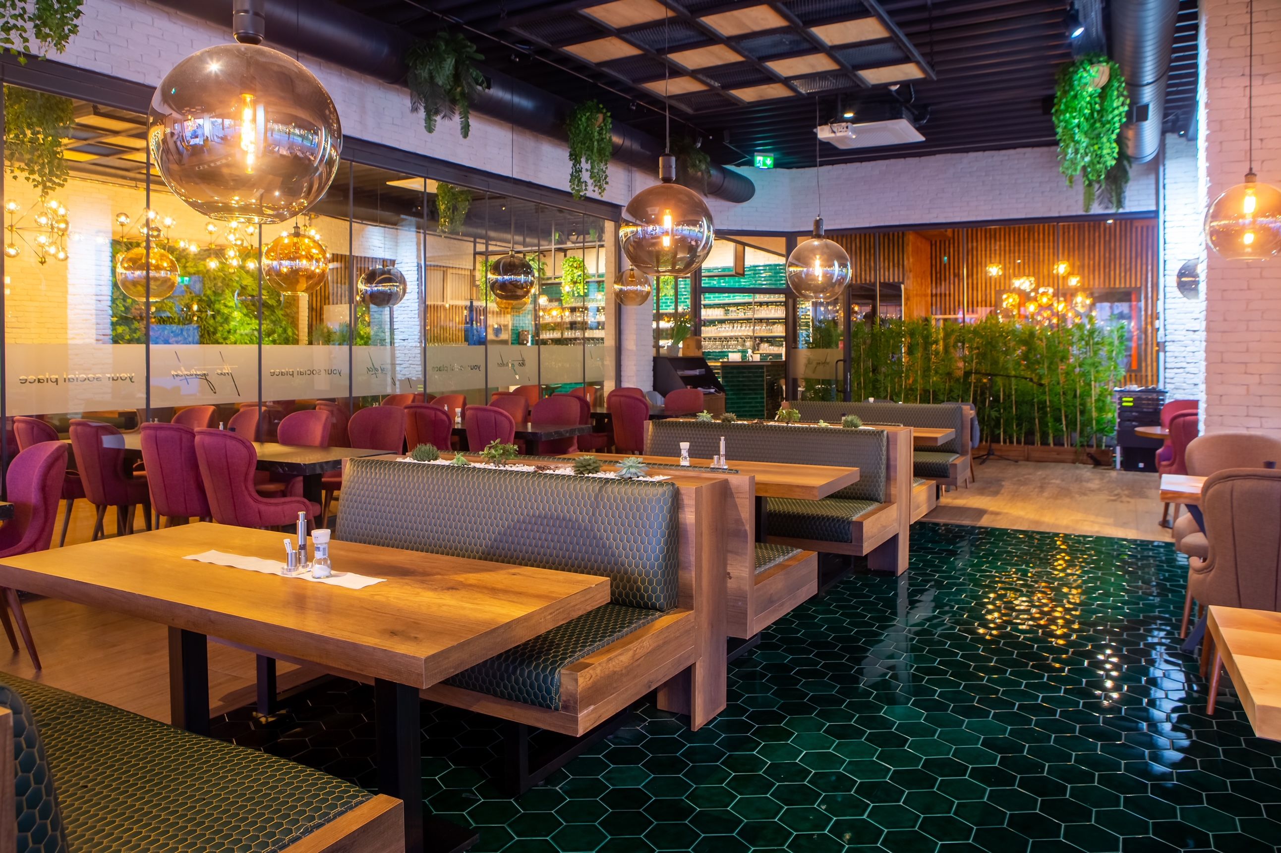 Innenansicht eines modernen Restaurants mit Holztischen, grünen Fliesen und hängenden Pflanzen.