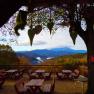 Terrace with wooden tables and benches, surrounded by trees, with a view of wooded mountains and clouds on the horizon.