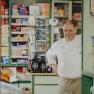 A man in chef's clothes stands smiling in an old store with shelves full of products.