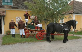 Historische Feuerwehrkutsche mit Pferd und Feuerwehrleuten in Uniform vor einem gelben Gebäude.