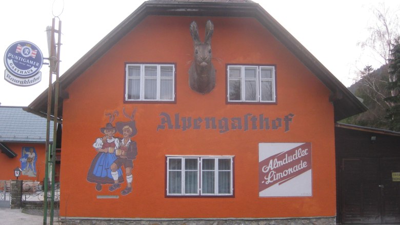 Orange gestrichenes Gebäude mit der Aufschrift 'Alpengasthof', Wandmalerei von tanzendem Paar und Almdudler-Werbung.