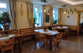 Innenansicht eines gemütlichen Gastraums mit Holztischen und Stühlen, dekoriert mit Pflanzen und Hirschgeweihen an der Wand.