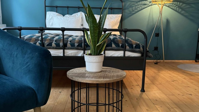 Schlafzimmer mit blauem Sessel, Metallbett, Pflanze auf Tisch und Stehlampe.