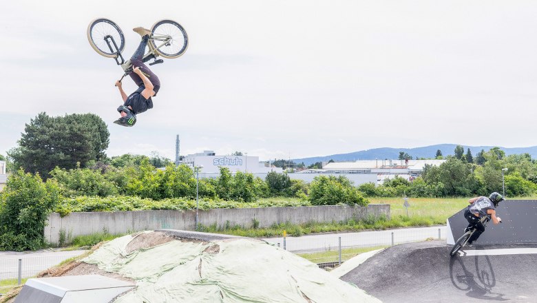 Zwei BMX-Fahrer auf einer Pumptrack-Anlage, einer macht einen Rückwärtssalto, der andere fährt eine Kurve.