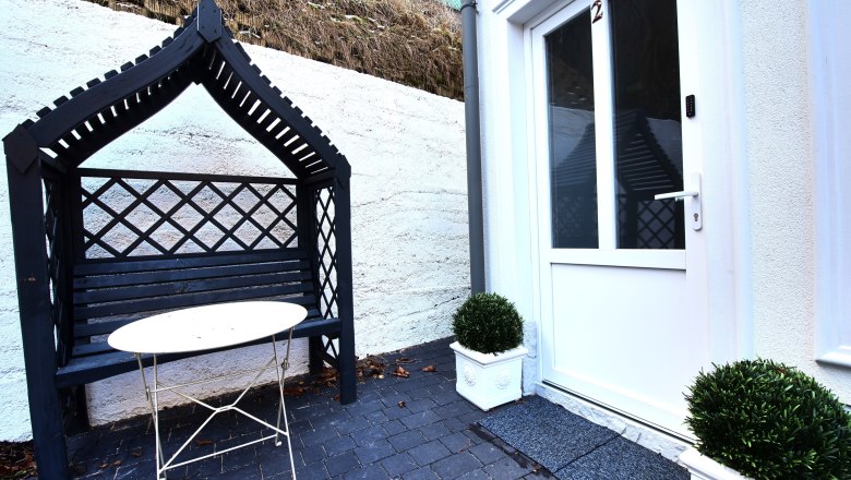 Eingangsbereich mit weißer Tür, schwarzer Bank mit Überdachung und kleinem Tisch.