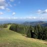 Blick auf eine gr&uuml;ne Berglandschaft mit W&auml;ldern und blauem Himmel.