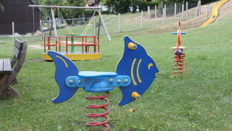 Spielplatz mit Wipptier in Fischform, Karussell und Rutsche.