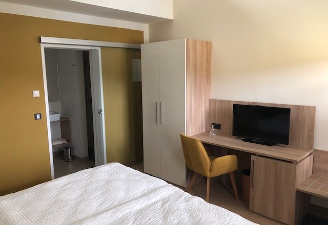 Ein modernes Gästezimmer mit Bett, Schreibtisch, Stuhl, Fernseher und Schrank. Im Hintergrund ist ein Badezimmer zu sehen.