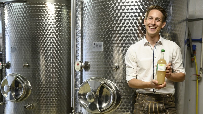 Mann in traditioneller Kleidung hält eine Flasche vor großen Metalltanks.