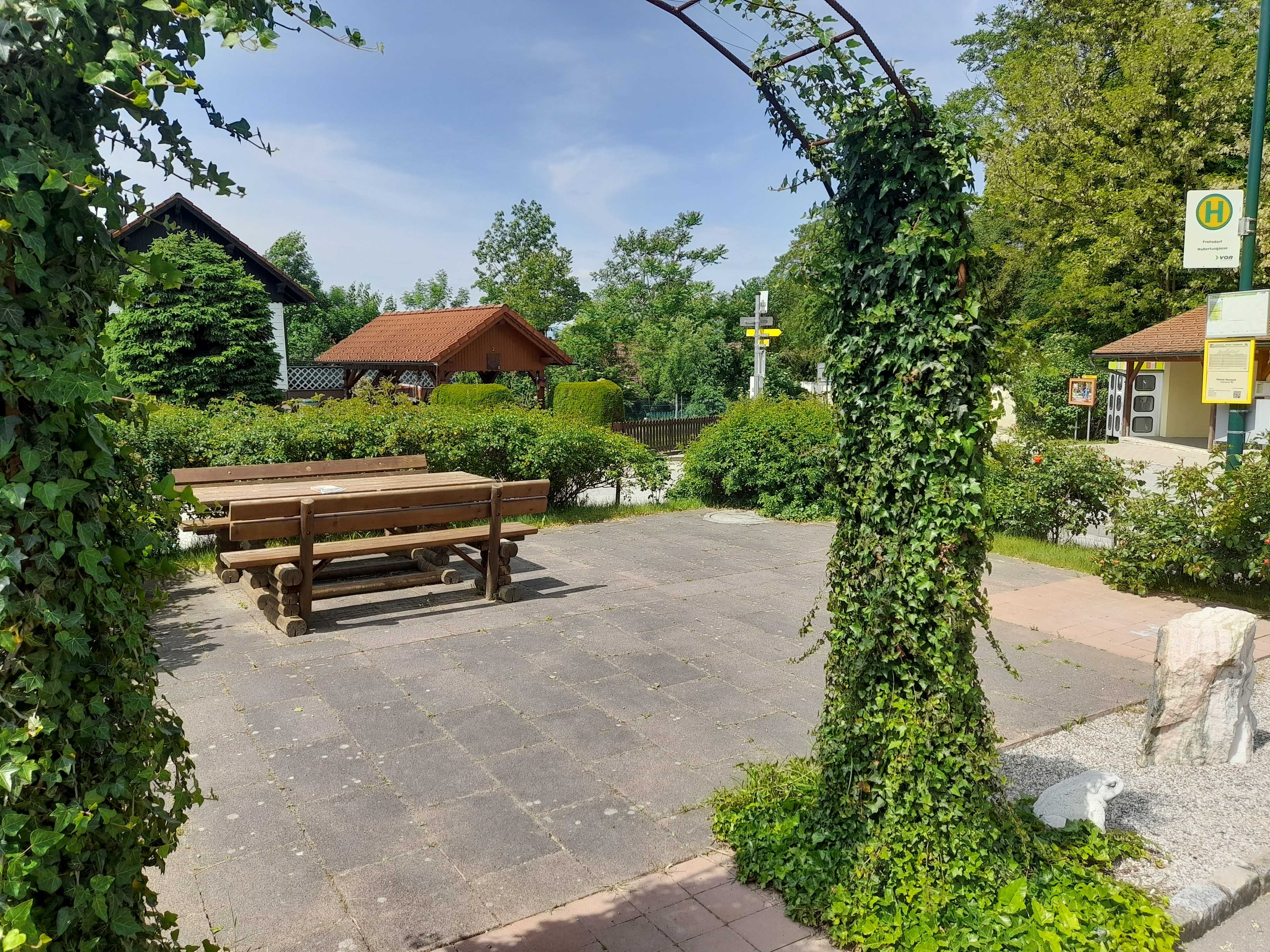 Ein ruhiger Platz mit Holzbänken und Tischen, umgeben von grünen Pflanzen und einem mit Efeu bewachsenen Torbogen.