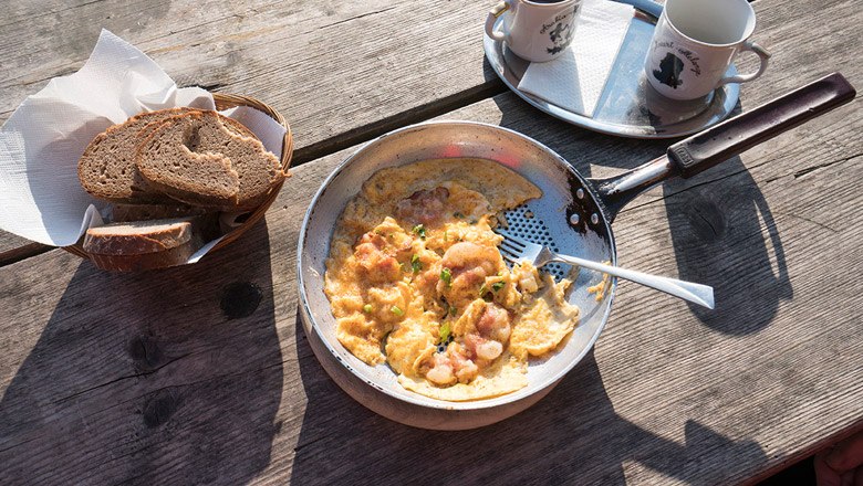 Pfanne mit Rührei und Speck auf Holztisch, daneben Brotkorb und zwei Tassen.