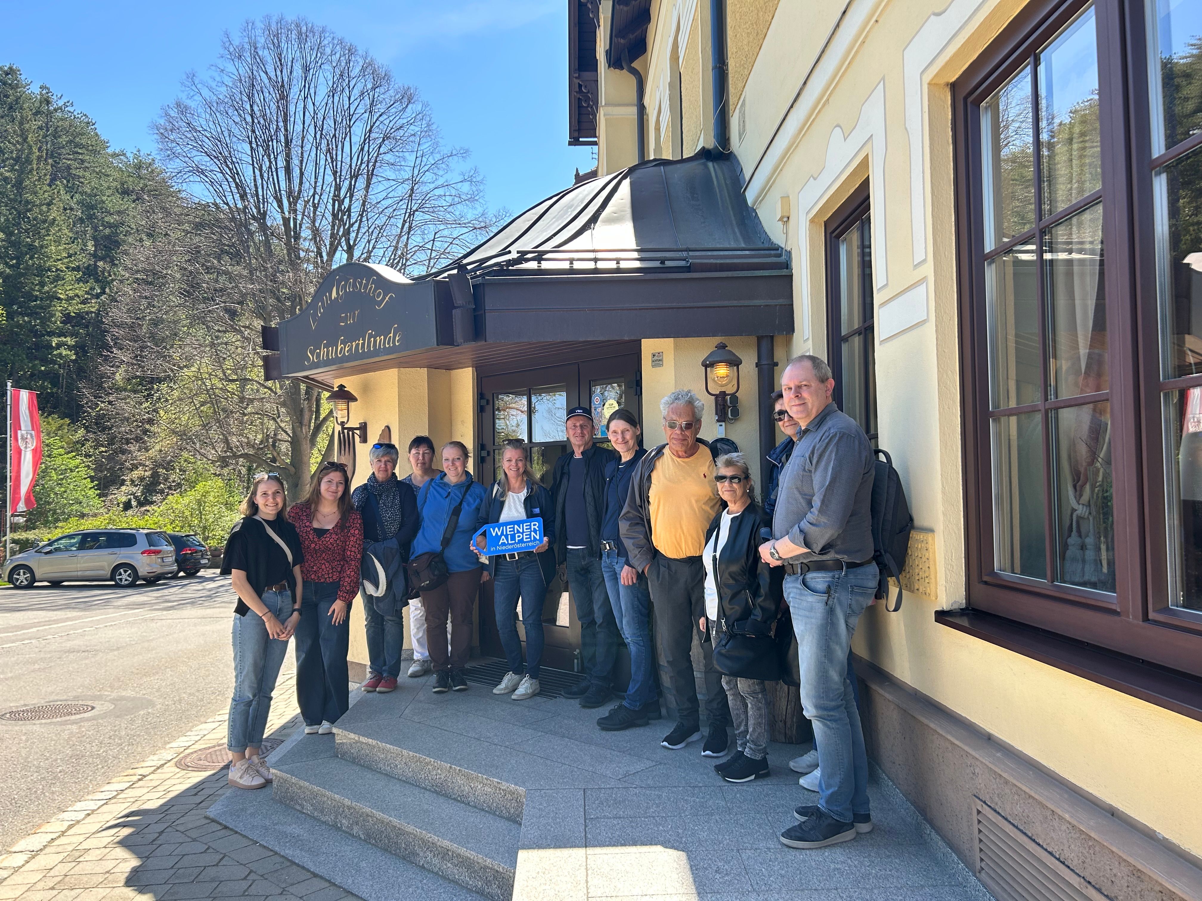 Eine Gruppe von rund 12 Personen steht auf den Stufen vor dem "Landgasthof zur Schubertlinde". Eine Frau hält ein Wiener Alpen Logo-Schild in der Hand.