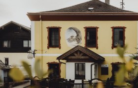 Gelbes Gebäude mit Wandgemälde eines Adlers und roten Fensterrahmen.