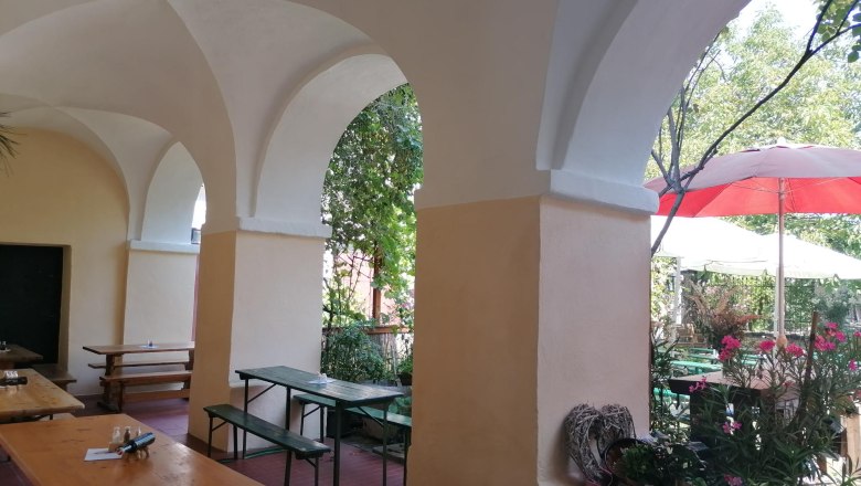 Arkaden mit Holzbänken und Tischen, Pflanzen und Sonnenschirmen im Hintergrund.