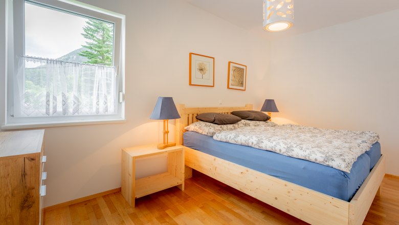 Ein gemütliches Schlafzimmer mit einem Holzbett, blauer Bettwäsche und zwei Nachttischlampen. Ein Fenster mit Blick auf Bäume und Berge.