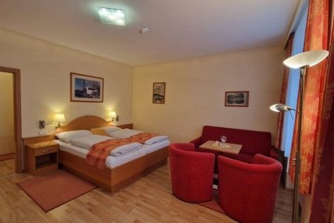 Hotelzimmer mit Doppelbett, rotem Sofa und Sesseln, Holzboden und Wandbildern.