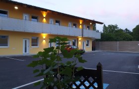 Außenansicht eines zweistöckigen Gebäudes mit gelber Fassade und beleuchtetem Balkon, davor ein geparkter roter Wagen und Pflanzen im Vordergrund.