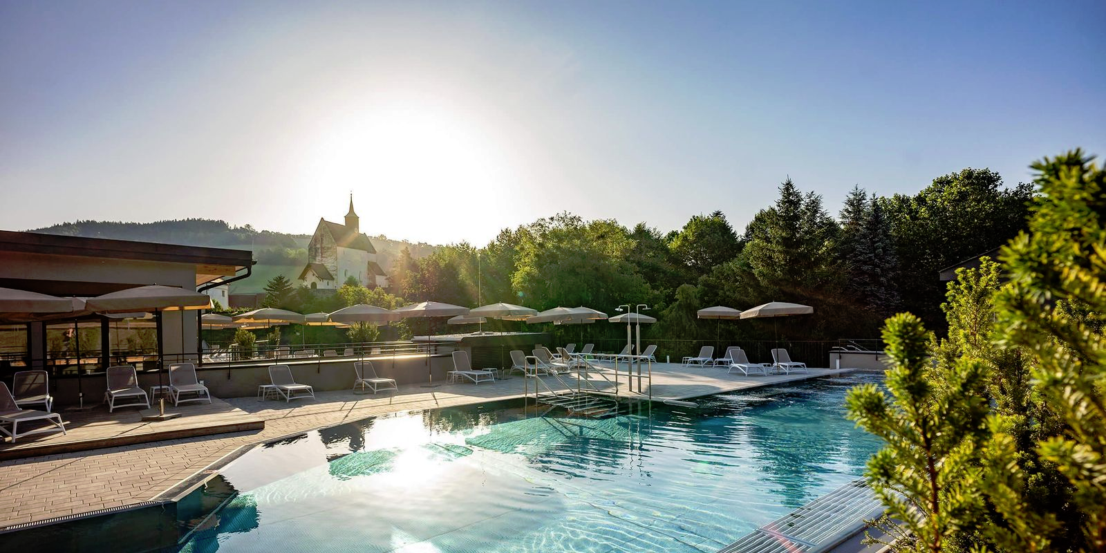 Außenanlage eines Hotels mit Pool, Liegestühlen und Schirmen, umgeben von Bäumen. 