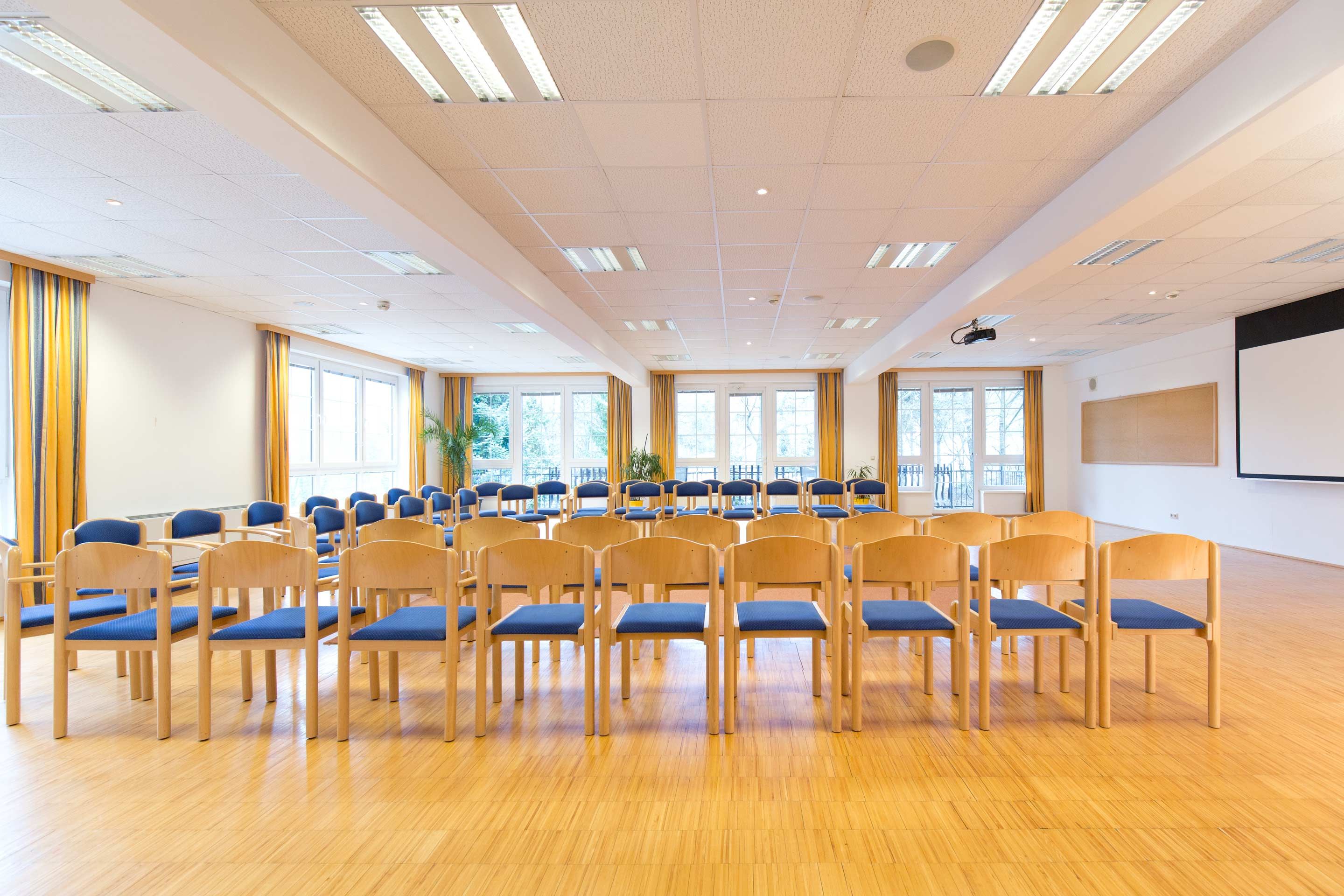 Ein heller Seminarraum mit Stuhlreihen, großen Fenstern und einem Projektor an der Decke.