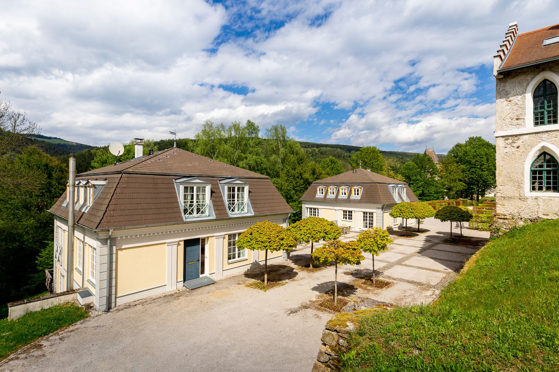 Zwei Kavaliershäuser mit Mansardendächern und gepflegten Bäumen im Vordergrund, umgeben von grüner Landschaft und blauem Himmel.