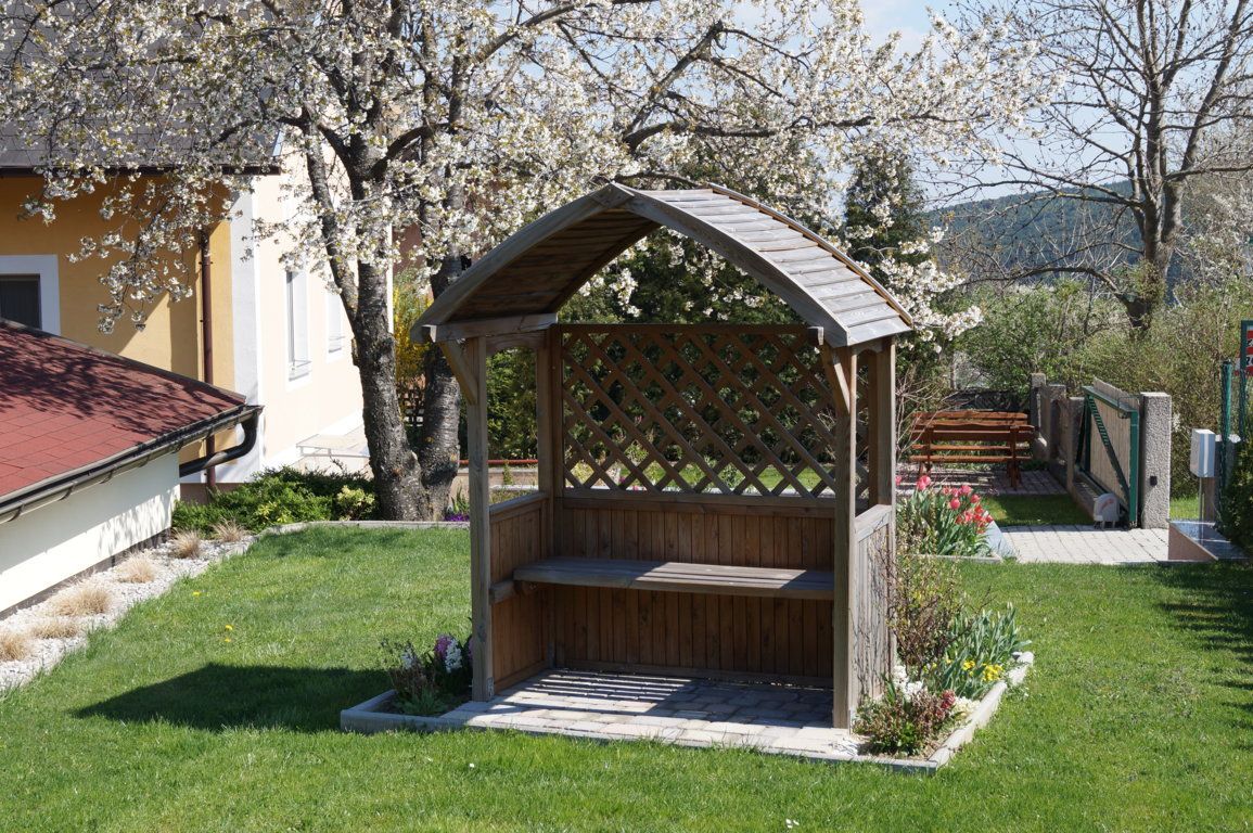 Ein Holzbankerl mit Überdachung steht in einem blühenden Vorgarten neben einem blühenden Baum.