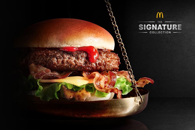 Ein Burger mit Salat, Käse, Speck und Ketchup auf einer Waage, daneben das McDonald's Logo und der Schriftzug 'The Signature Collection'.