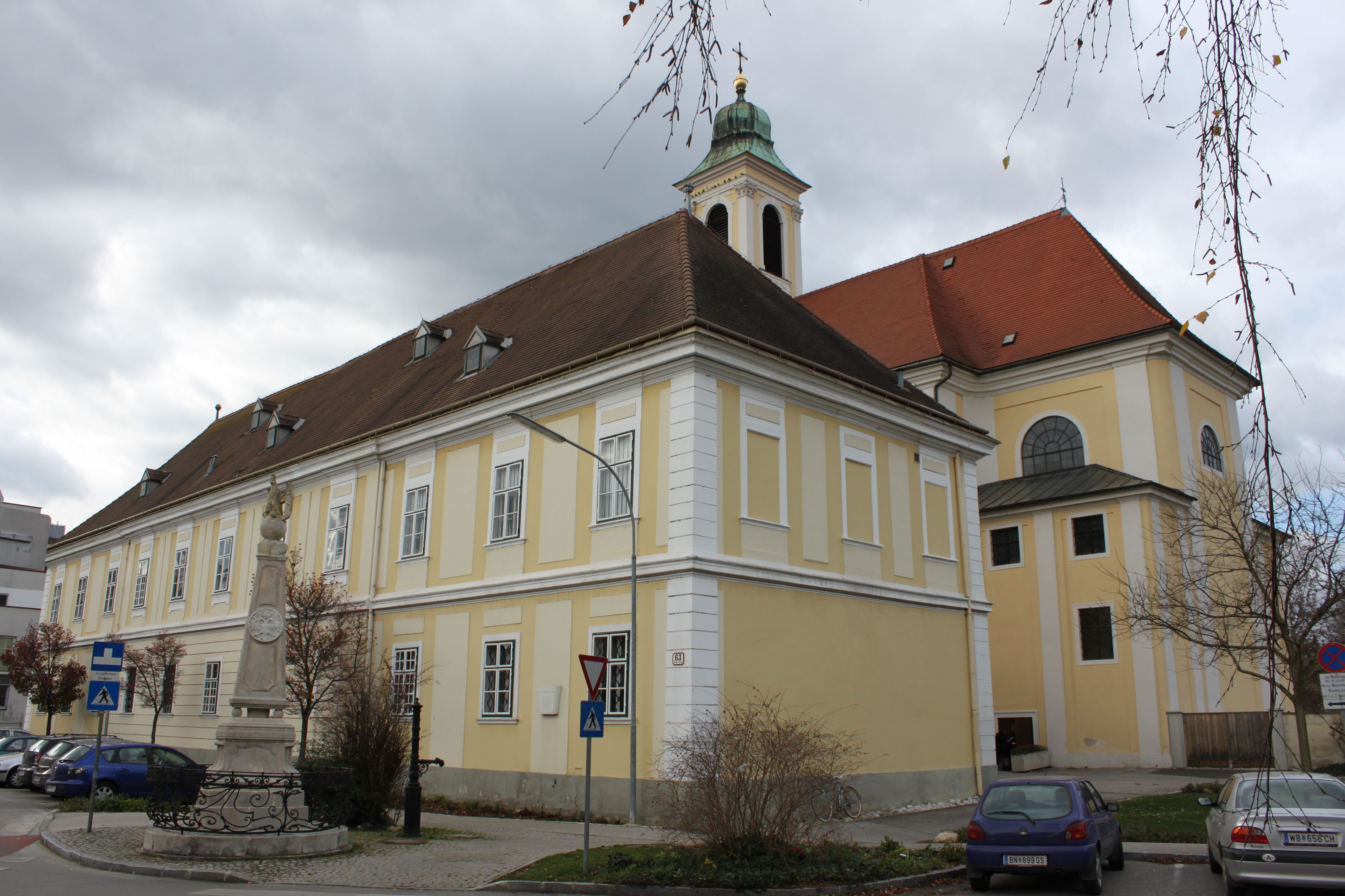 Historisches Gebäude mit gelber Fassade und rotem Dach, daneben eine Statue und parkende Autos.