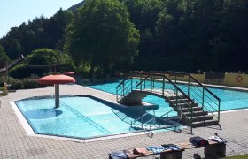 Ein Freibad mit einem kleinen Pool, einer Br&uuml;cke und einem Pilz-Wasserspiel. Umgeben von B&auml;umen und einem sonnigen Himmel.