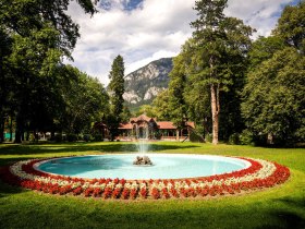 Inmitten einer malerischen Landschaft sprudelt der Brunnen in einem liebevoll gestalteten Garten, umgeben von bunten Blumenbeeten. Die majest&auml;tischen Berge im Hintergrund verleihen der Szenerie eine beeindruckende Kulisse, die zum Verweilen und Entspannen einl&auml;dt.