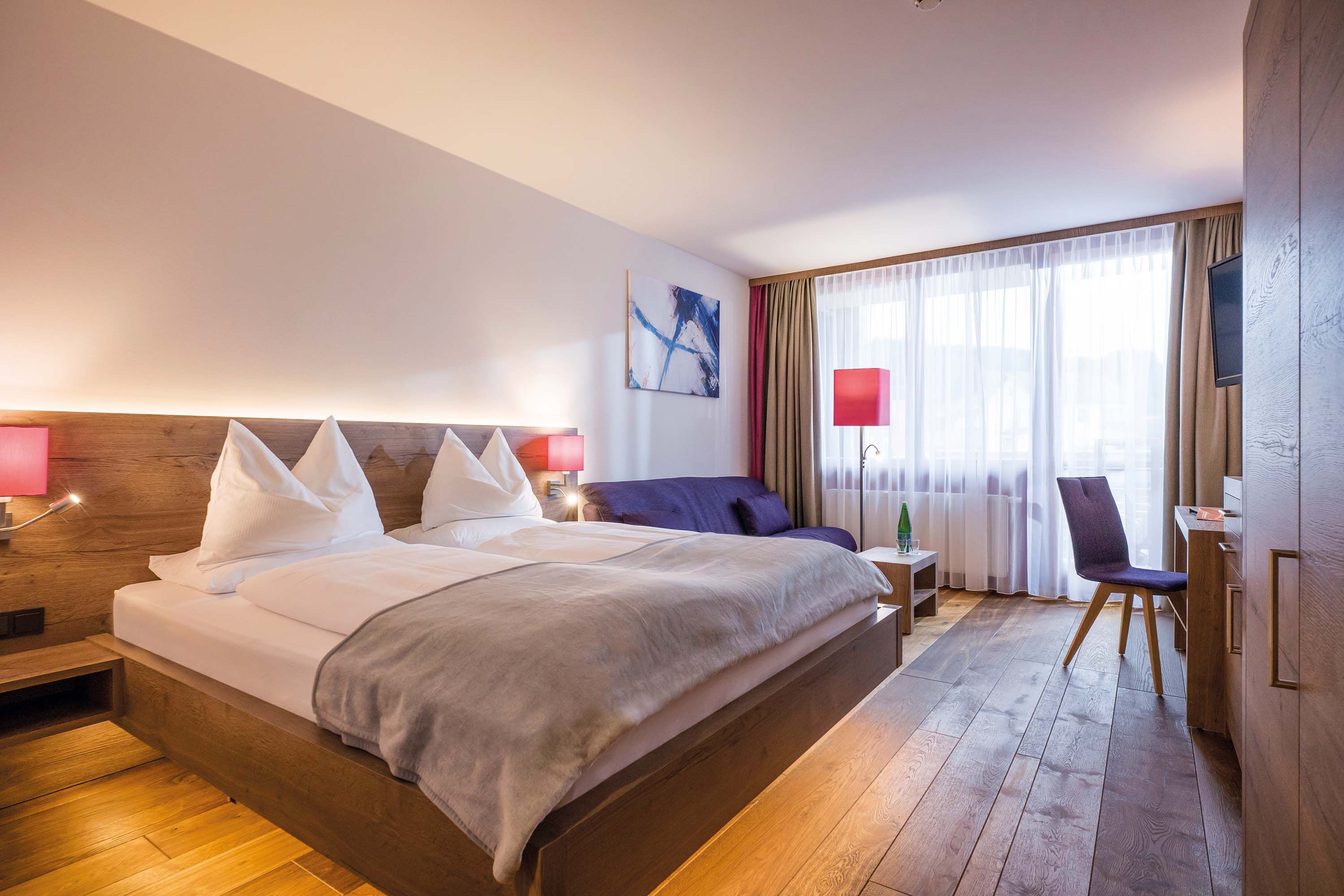 ein helles, modernes Hotelzimmer mit Doppelbett, Holzboden, Schreibtisch und Fensterwand