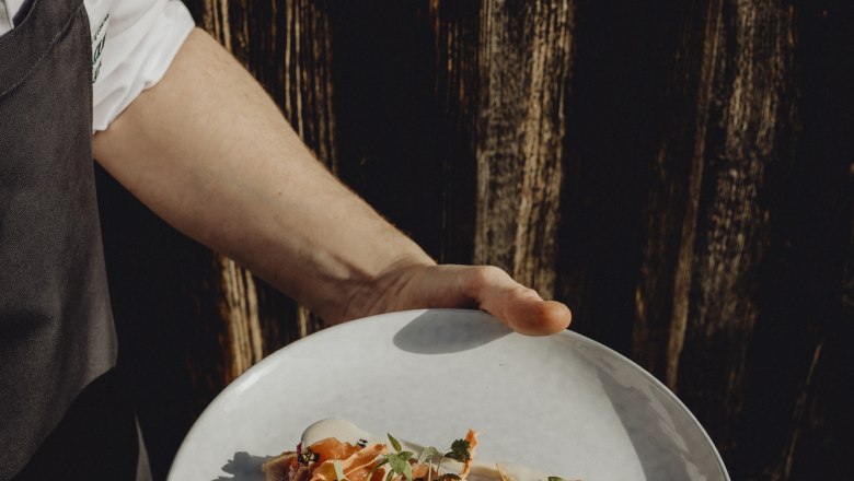 Egy tányér művészien elrendezett Vitello Forellóval, amelyet egy szakácsruhás személy tart a kezében egy sötét faháttér előtt.