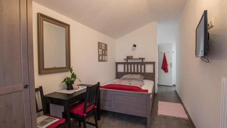 A cozy single room with wooden furniture, a bed, a small table with chairs, a mirror and a TV on the wall.