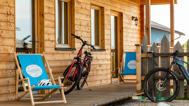 Wooden veranda with two bicycles and blue deckchairs in front of a wooden house.