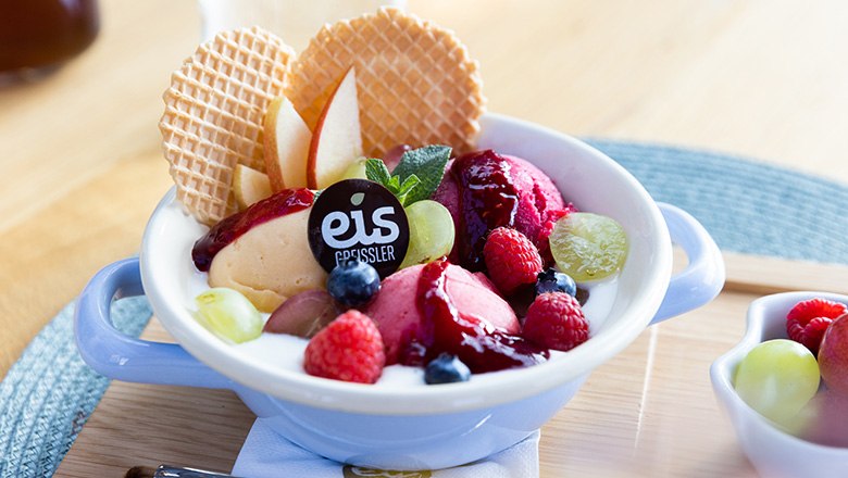 Eisbecher mit Joghurt, Fruchteis, Fr&uuml;chten und Waffeln auf einem Holzbrett.