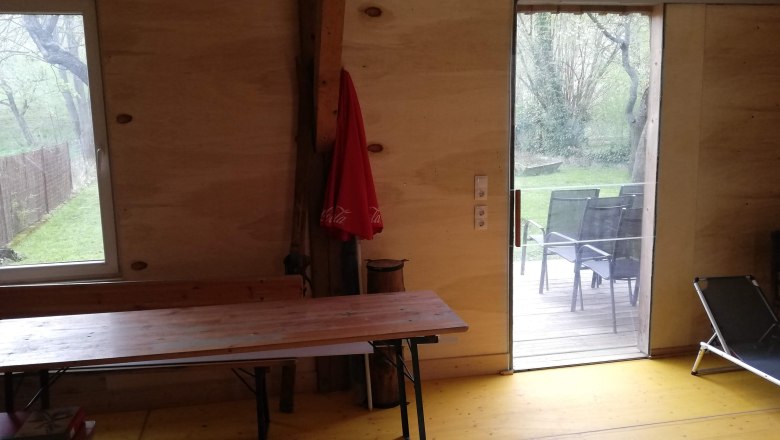 Innenansicht eines Raumes mit Holzwänden, Tisch, Liegestuhl und Blick auf Terrasse und Garten.