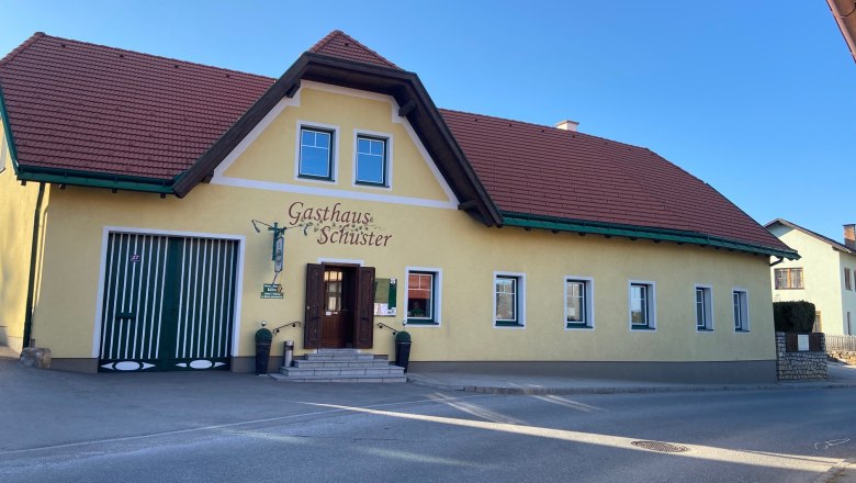 Gelbes Gebäude mit rotem Dach und der Aufschrift 'Gasthaus Schuster'.