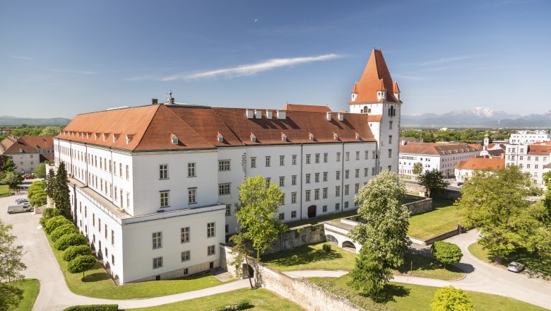 Blick auf eine historische Milit&auml;rakademie mit rotem Dach und Turm, umgeben von gr&uuml;nen B&auml;umen und Rasenfl&auml;chen, im Hintergrund Berge.