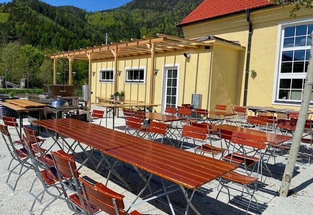 Biergarten mit Holztischen und Stühlen vor einem gelben Gebäude.
