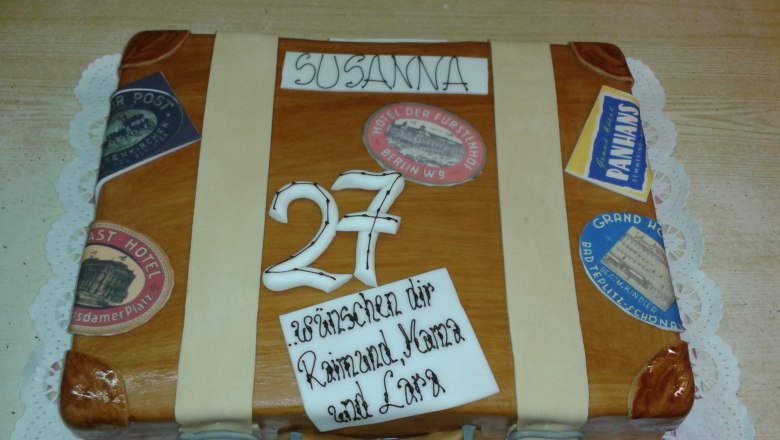 A cake in the shape of a suitcase with stickers and the number 27.