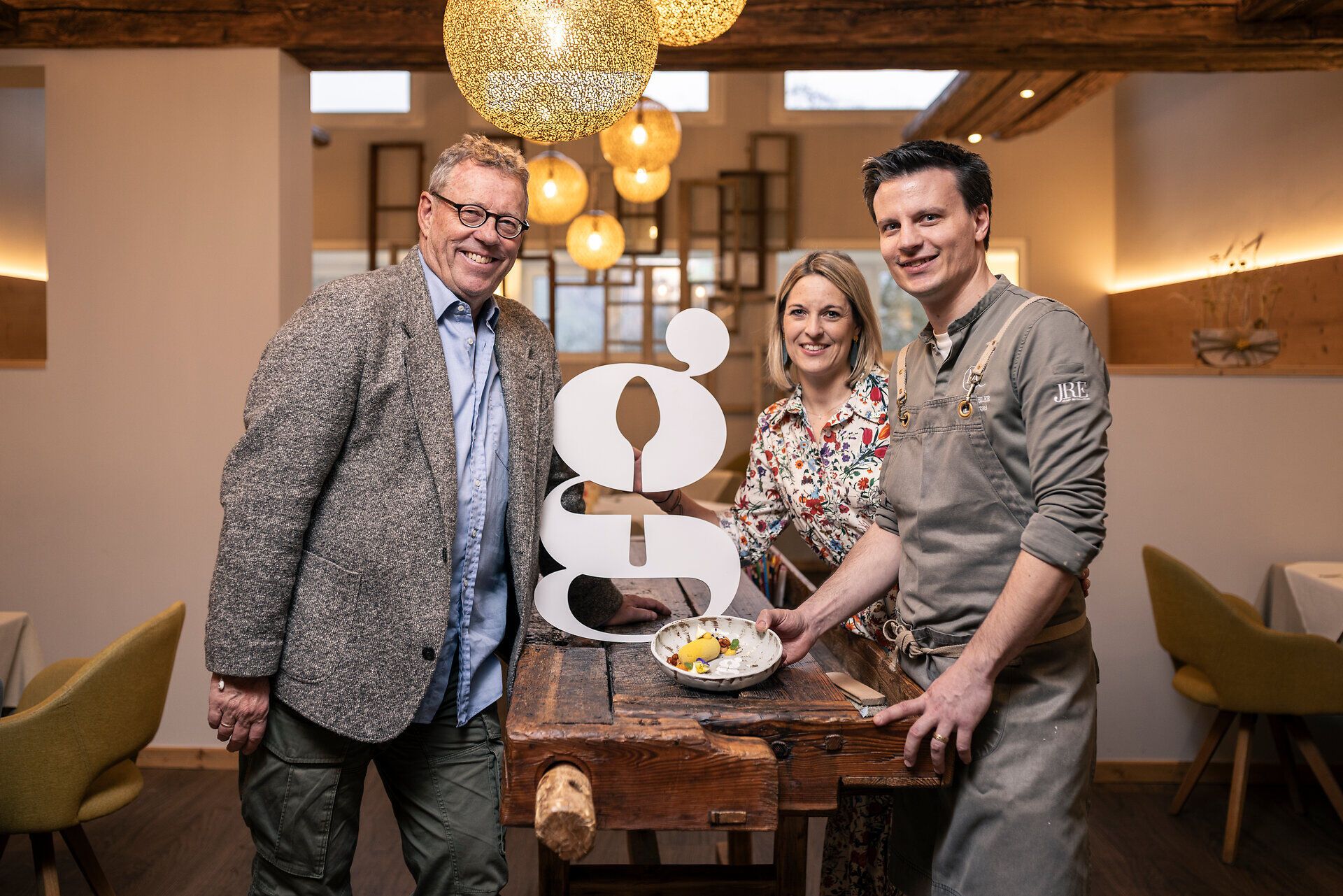 Herbert Steinböck mit Nina und Peter Pichler stehen rund um einen Holztisch. Darauf ist ein weißes kleiner G und ein angerichteter Teller platziert.