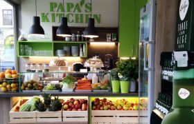 Innenansicht eines modernen Lokals mit frischem Obst und Gem&uuml;se, gr&uuml;ner Dekoration und dem Schriftzug 'Papa's Kitchen'.