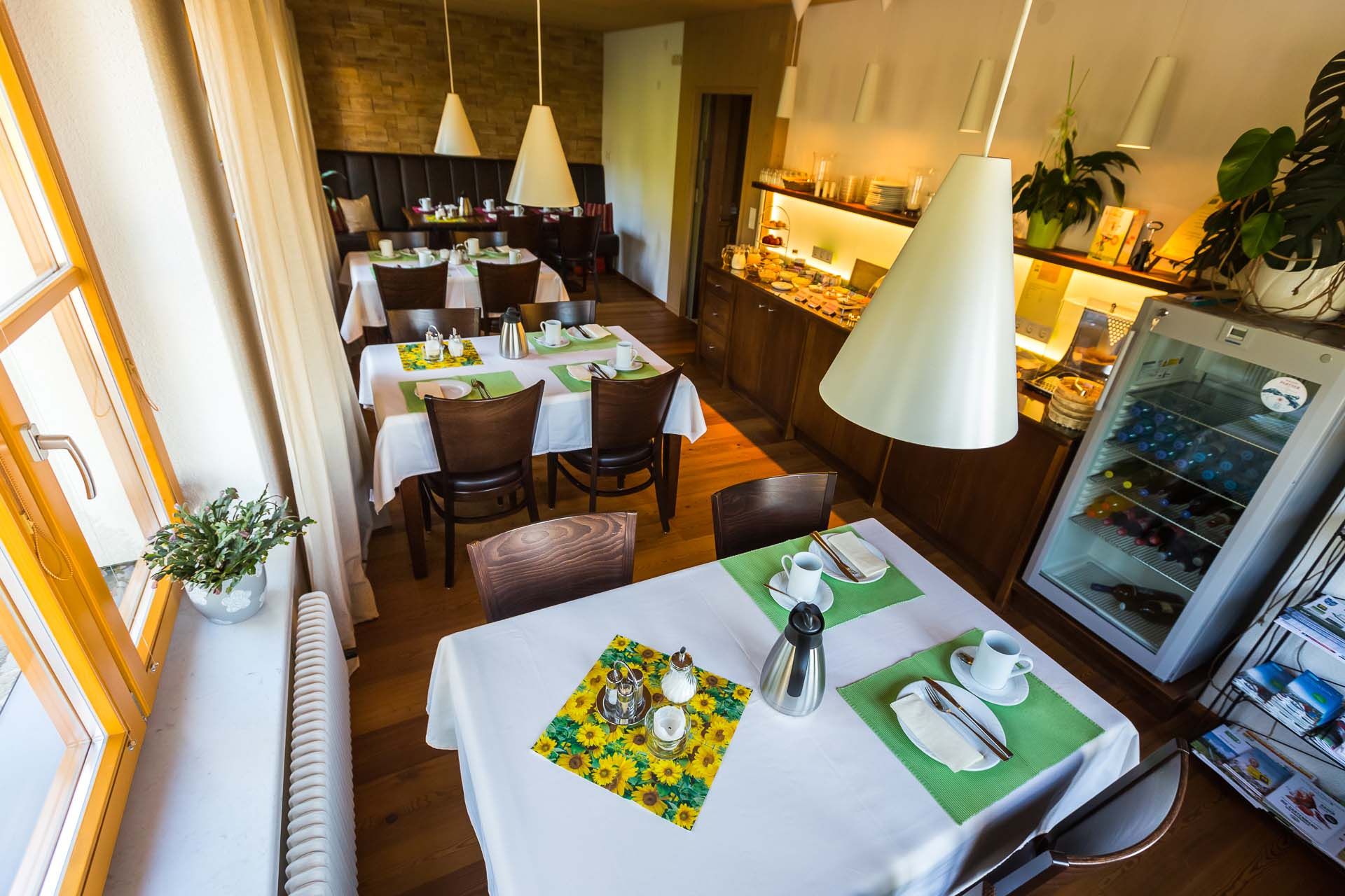 Ein gemütliches Frühstückszimmer mit gedeckten Tischen, weißen Tischdecken und grünen Platzsets. Im Hintergrund ein Buffet und ein Kühlschrank.