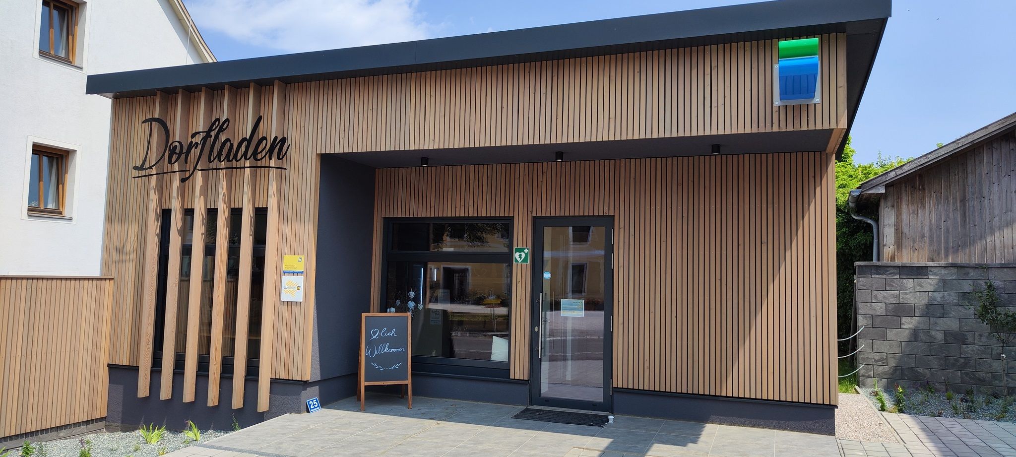 Außenansicht eines modernen Dorfladens mit Holzverkleidung und einem Willkommensschild vor der Tür.