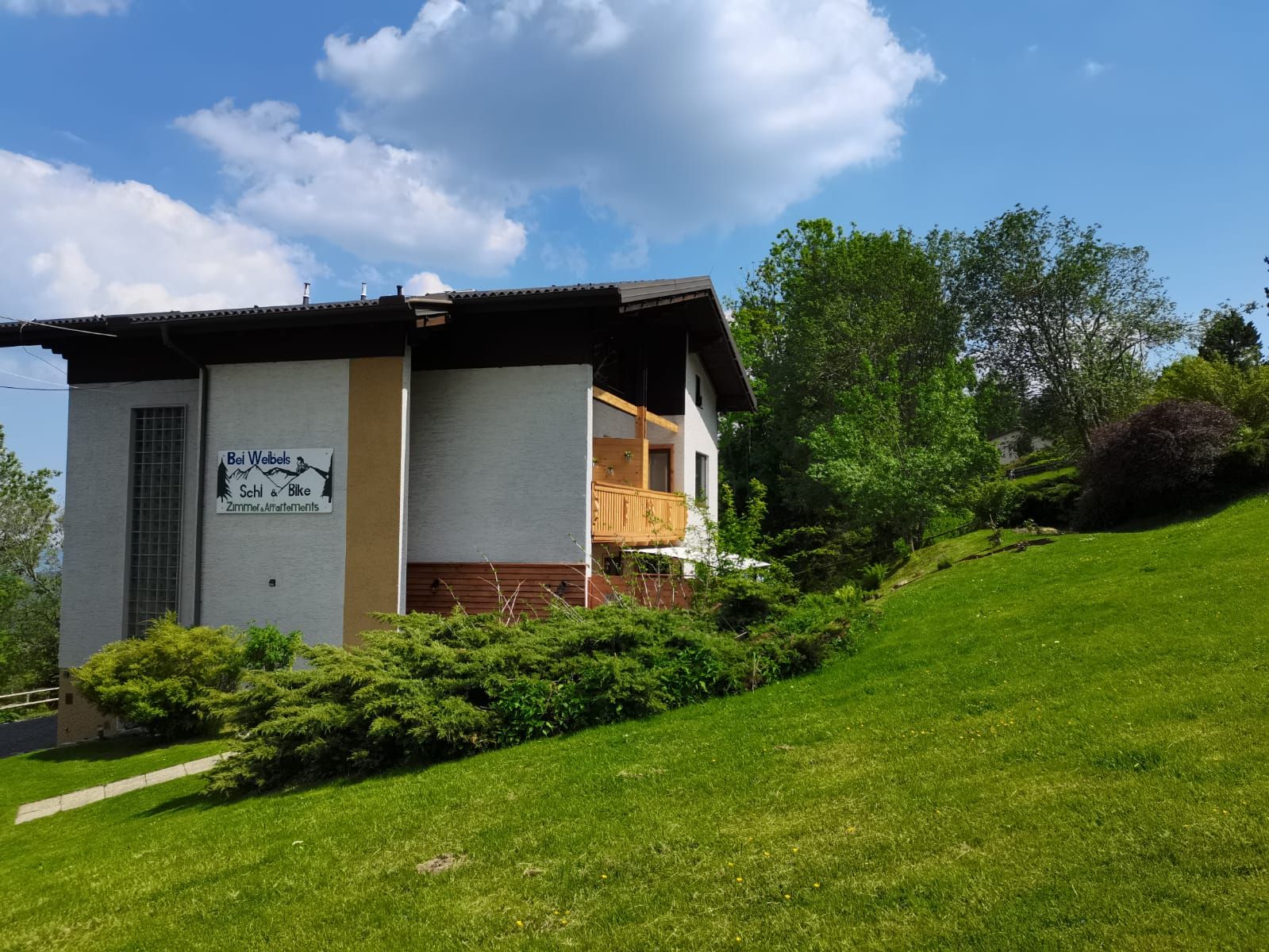 Haus mit Schild 'Bei Webels Ski & Bike' auf grüner Wiese, umgeben von Bäumen, unter blauem Himmel.
