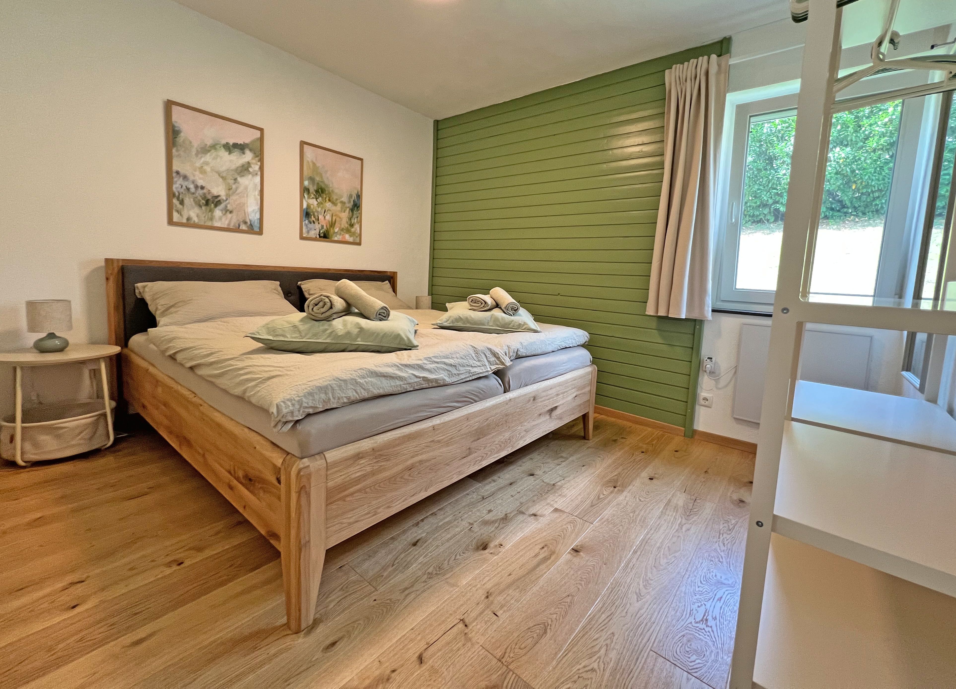 A modern bedroom with a double bed, a green accent wall, two pictures, a nightstand, and a window overlooking the greenery.
