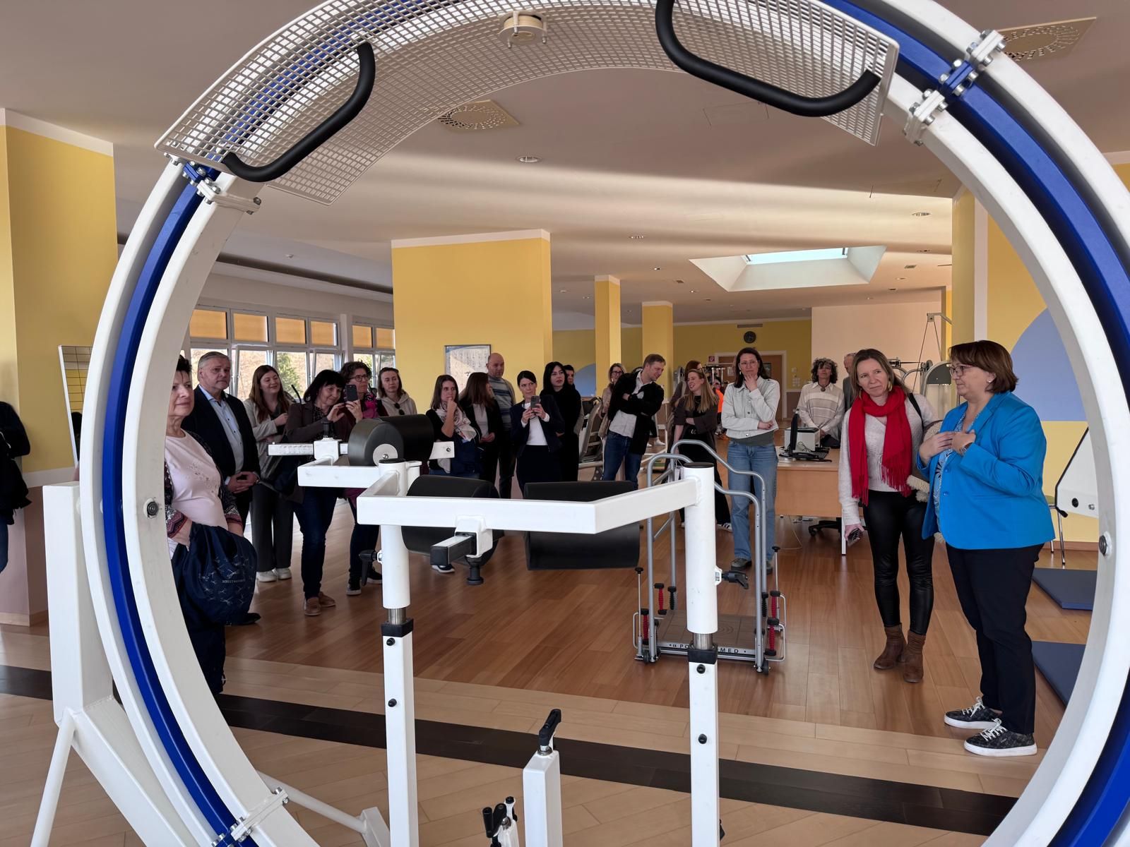 Etwa 20 Personen stehen in Alltagskleidung in einem großen Fitnessraum. 